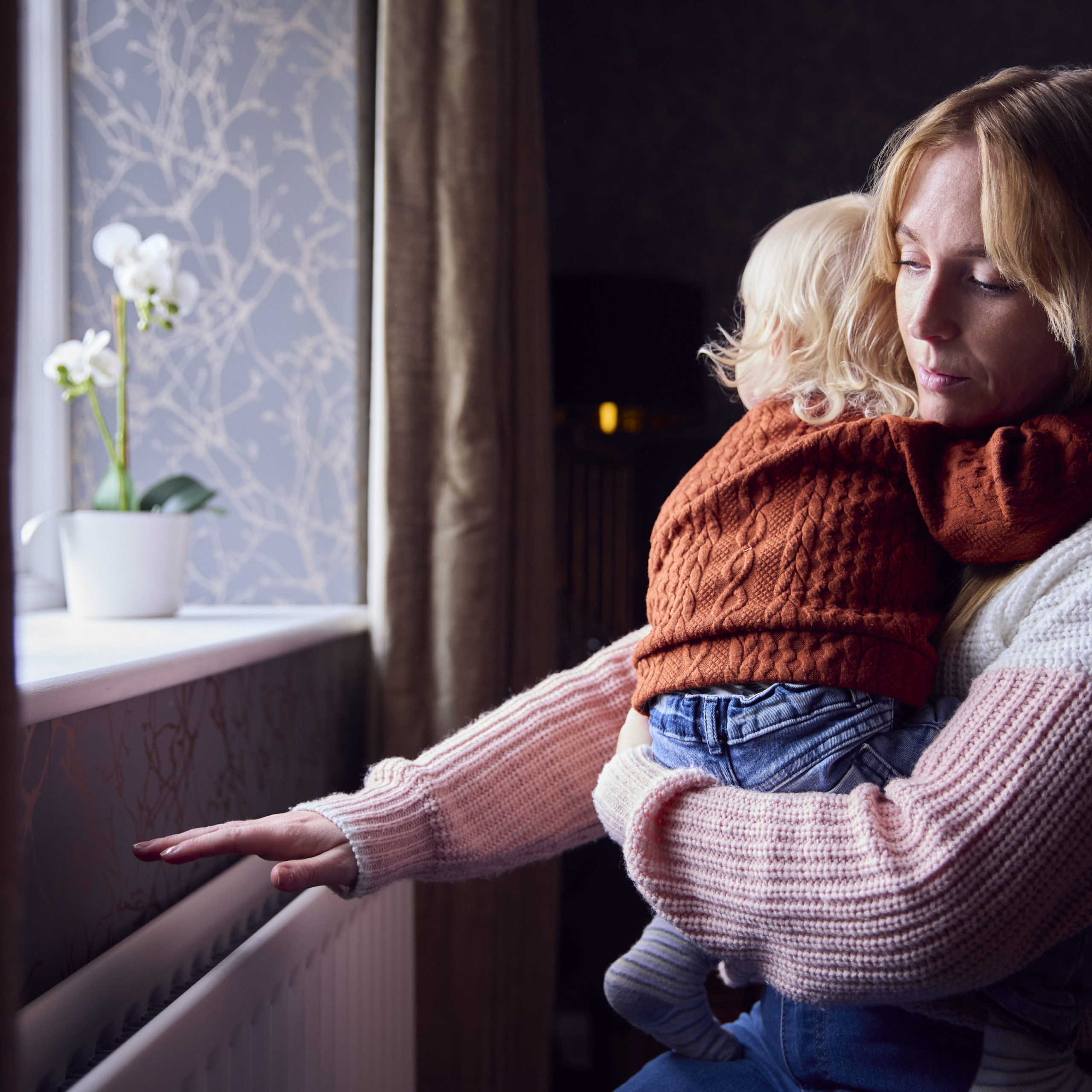 Mother carrying child in home whilst holding one hand over the radiator for warmth.
