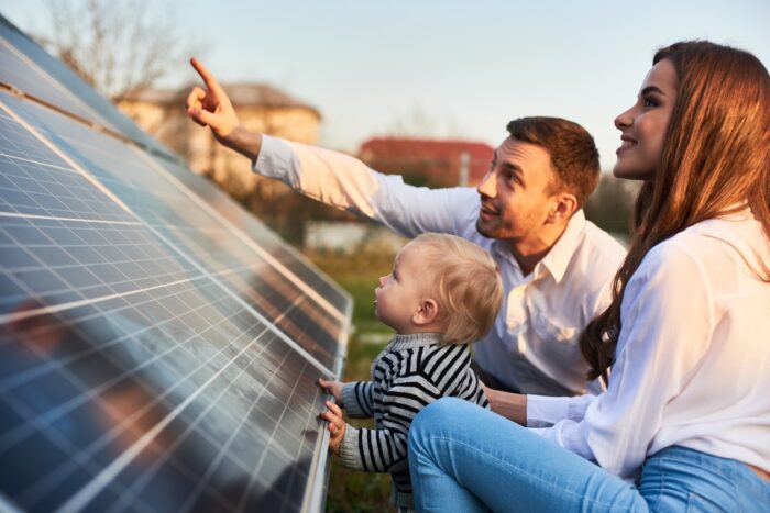 Man shows his family the solar panels on the plot near the house during a warm day. Young woman with a child and a man in the sun rays look at the solar panels.