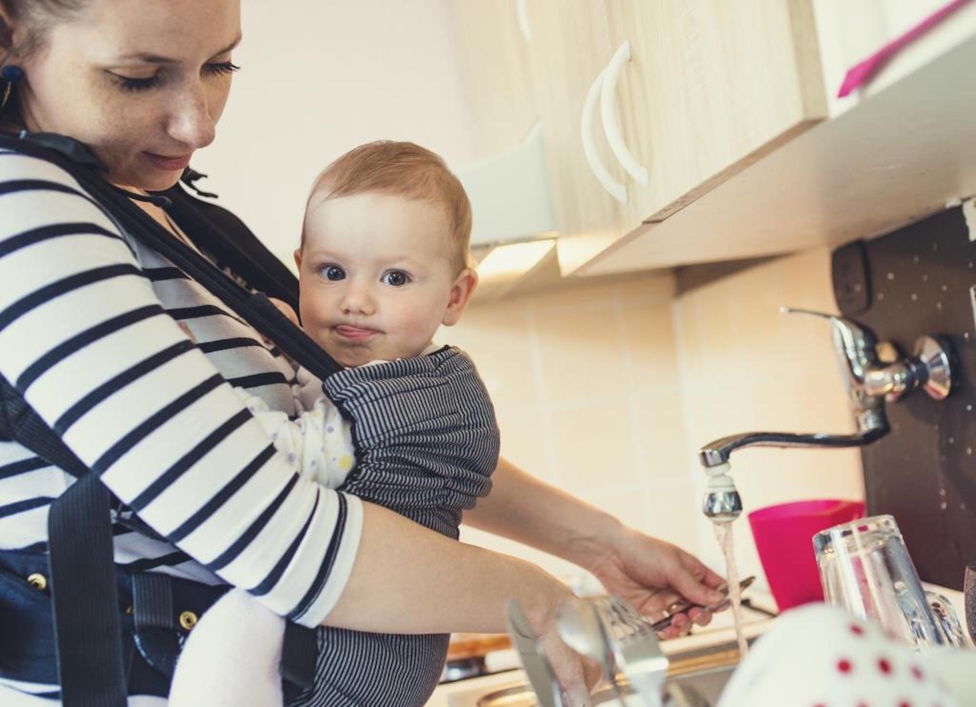 A mother in the kitchen doing the washing up whilst holding her young baby in a sling.