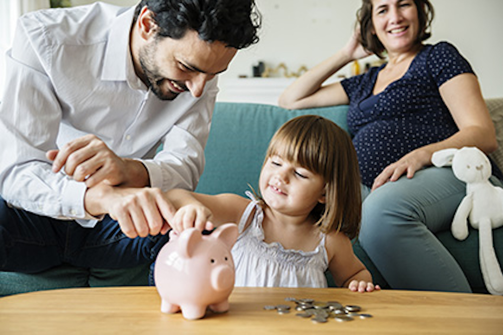 A family of three sitting on a sofa. The daughter is putting coins in a pink piggy bank