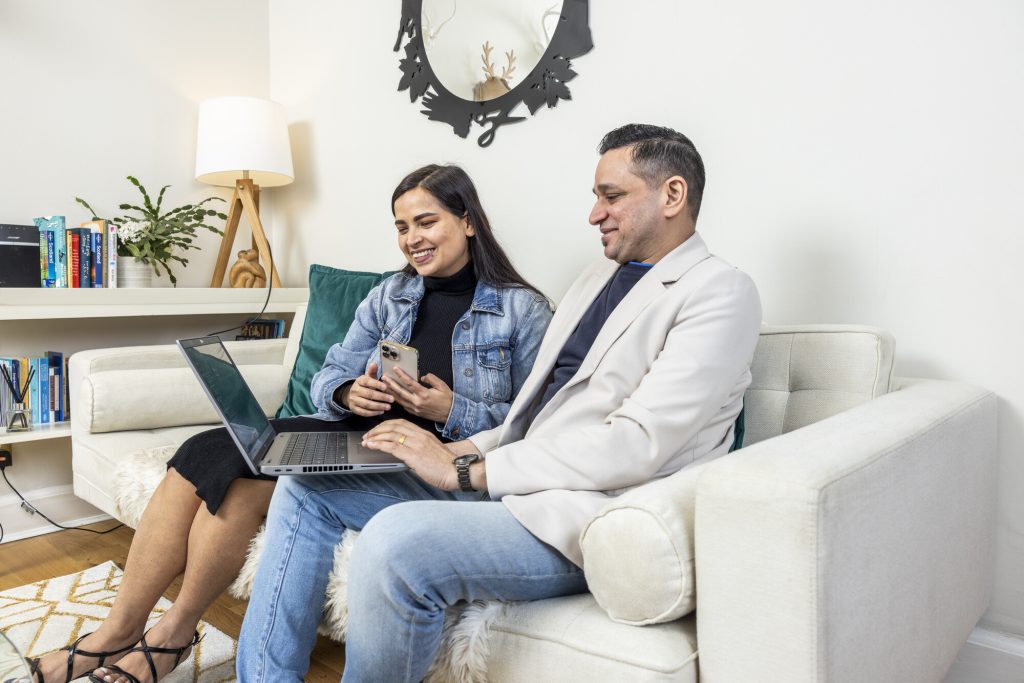 A male and female looking at their laptop whilst sat on the sofa.