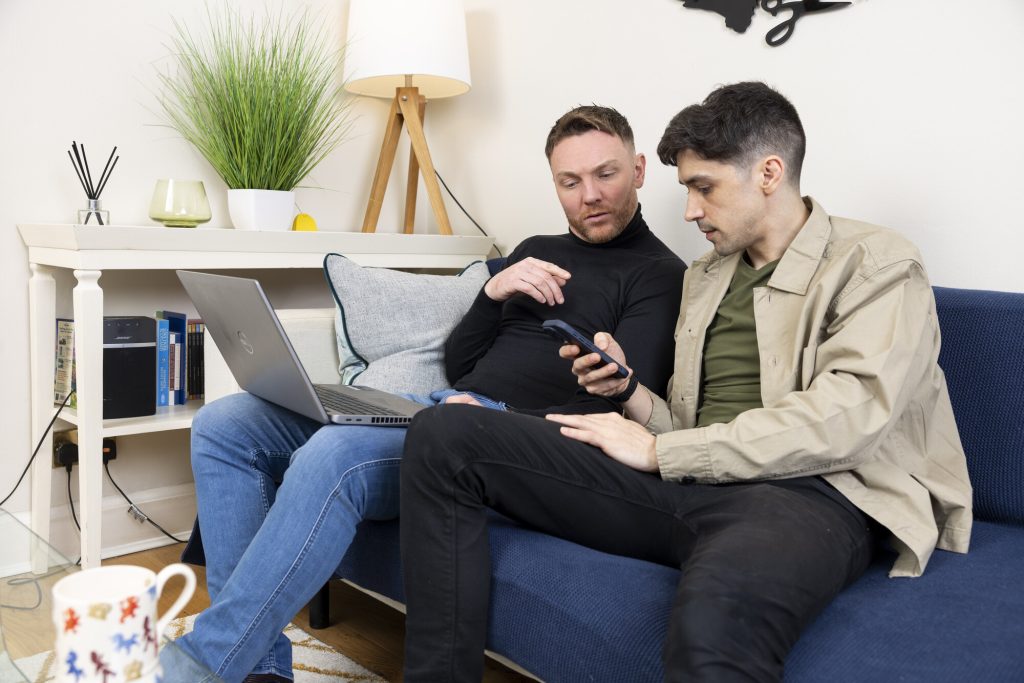 Two men sitting on a couch . Both are looking at a mobile phone, concerned about what they're viewing.