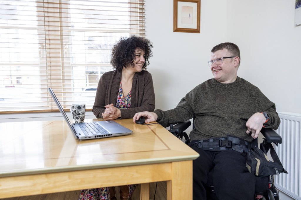 Female carer and male wheelchair user at a table, looking at a laptop